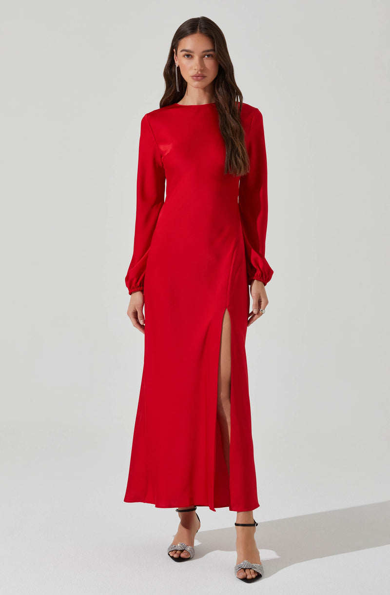 A full-length red evening gown featuring long sleeves with elasticized cuffs, a high round neckline, and a high slit on one side. The fabric appears smooth and slightly shiny, giving the dress an elegant look. The model is wearing open-toe heels with ankle straps.