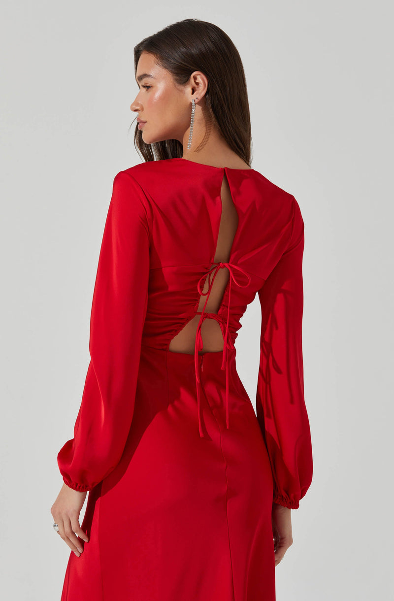 A woman is wearing a long, elegant red dress with long, puffed sleeves that are gathered at the wrists. The back of the dress features a large slit with a tie detail that creates an open back design. The dress is made of a shiny, satin-like fabric. She is accessorized with dangling, sparkling earrings.