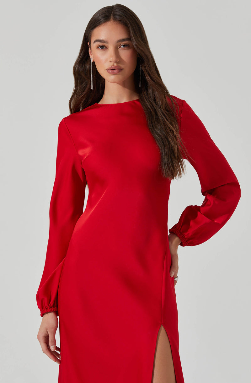 A woman is wearing a long, red satin dress with long sleeves and elasticized cuffs. The dress has a high, rounded neckline and features a high slit on one side. She is accessorized with long, dangling earrings.