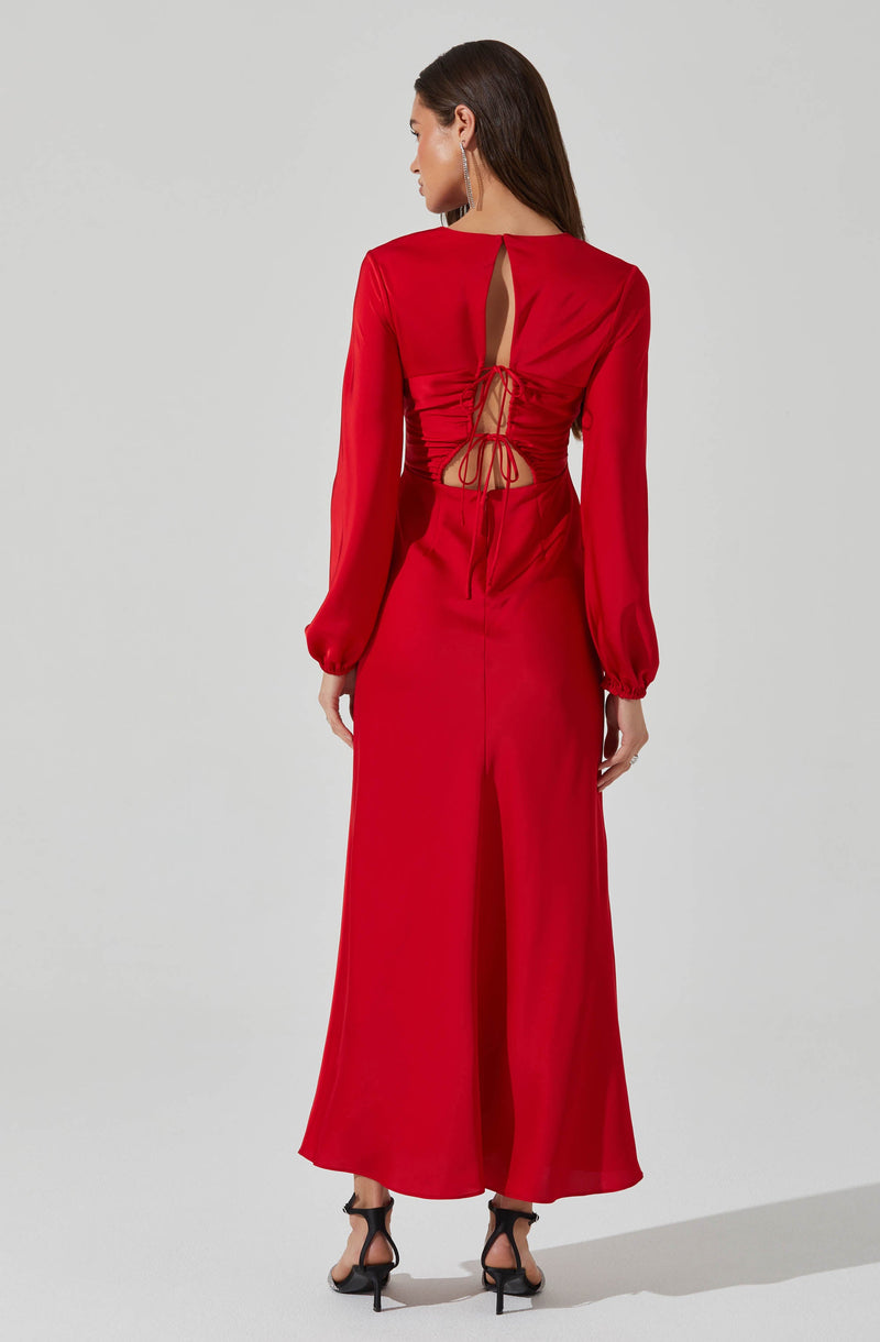 A woman is wearing a long, bright red dress with long sheer sleeves that are gathered at the wrists with elastic. The back of the dress features a keyhole opening at the upper back, and a ruched bodice with a lace-up tie detail at the waist, revealing some skin. The dress has a floor-length, flowing skirt. She is also wearing black high-heeled sandals with ankle straps.