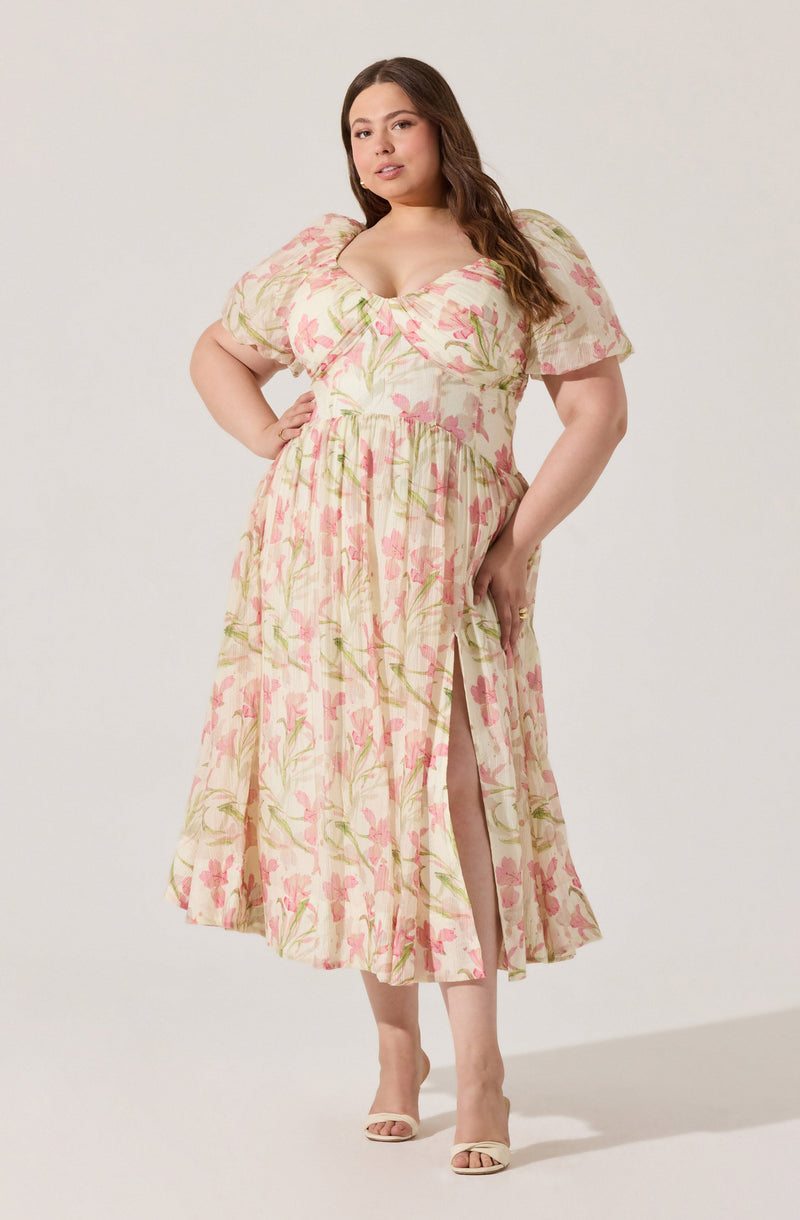 A woman is wearing a long, flowy dress with a light, cream-colored background and a pink and green floral pattern. The dress has short, puffed sleeves, a fitted bodice with a sweetheart neckline, and a high slit in the skirt that reveals her leg. She is also wearing open-toe heels.