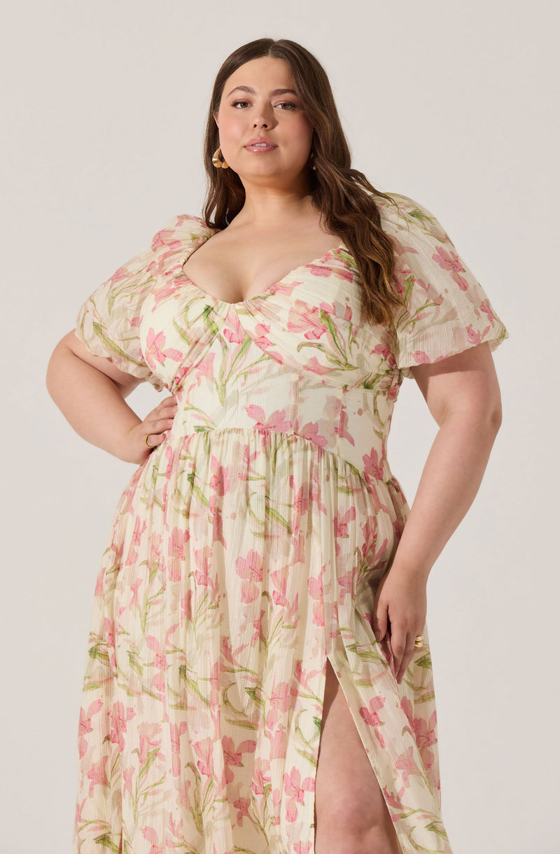 A woman is wearing a cream-colored maxi dress with a pink and green floral pattern. The dress features short, puffed sleeves, a v-neckline, and a tiered skirt with a high slit on one side.