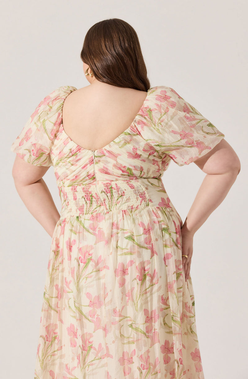 A woman is wearing a flowy dress with a pastel floral pattern featuring pink and green colors on a light, slightly transparent fabric. The dress has a deep, rounded back neckline and puffed short sleeves. The waist is cinched with a gathered detail, creating a fitted appearance around the midsection.