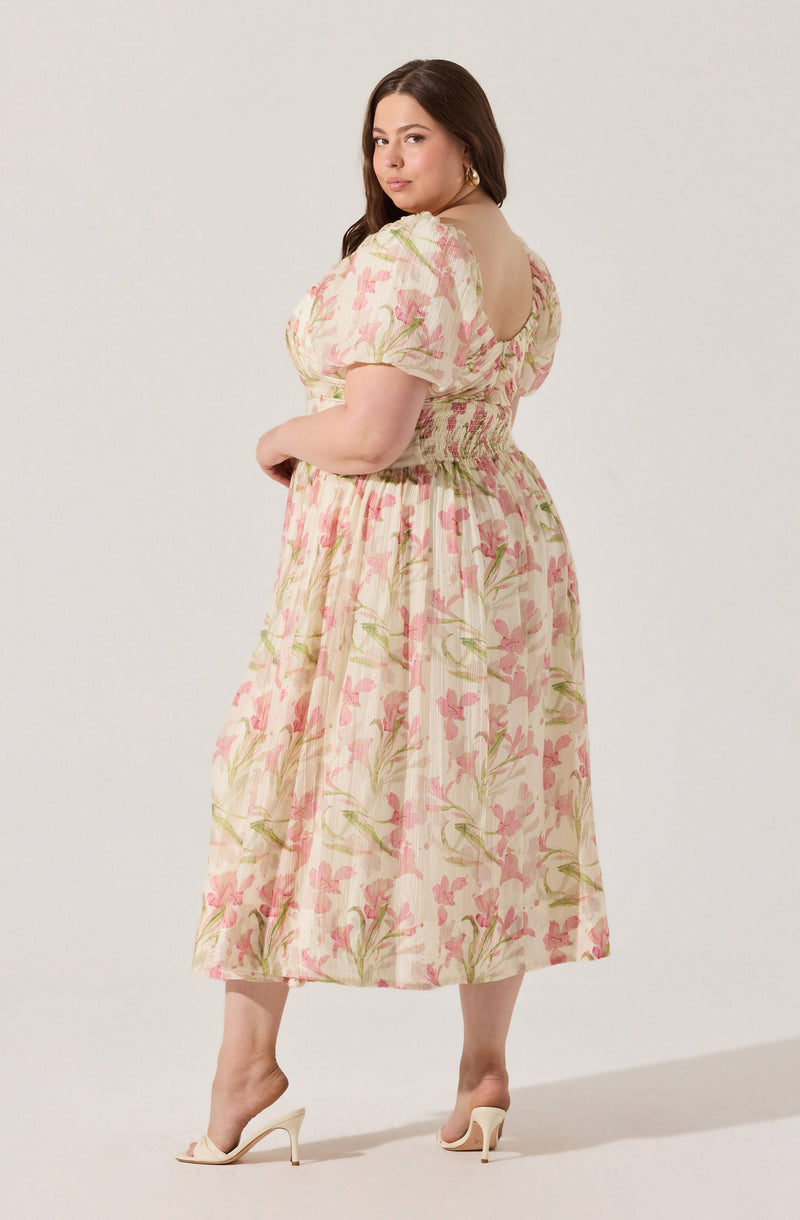 A woman is wearing a midi-length dress with a floral pattern consisting of pink flowers and green leaves on a light background. The dress has short, puffed sleeves and an elastic waistband. She is also wearing white high-heeled shoes.