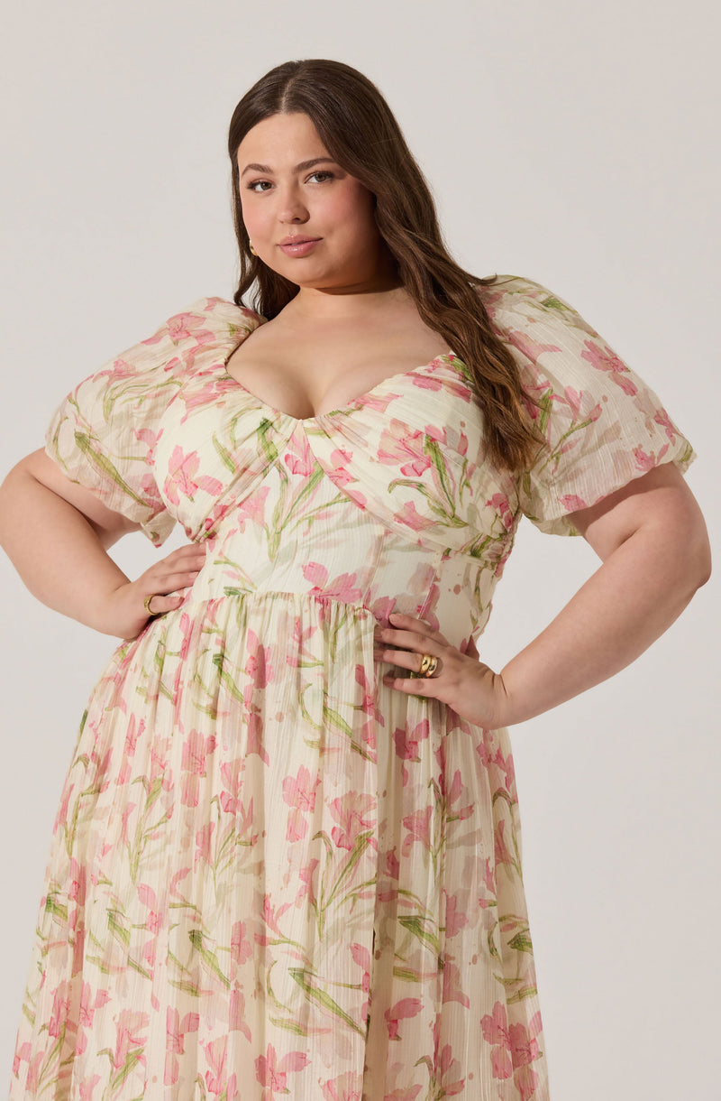 A woman is wearing a flowy, cream-colored dress with pink and green floral patterns. The dress has short, puffed sleeves and a fitted bodice that accentuates her waist, with a gathered skirt that falls to her ankles. The fabric appears lightweight and slightly textured.