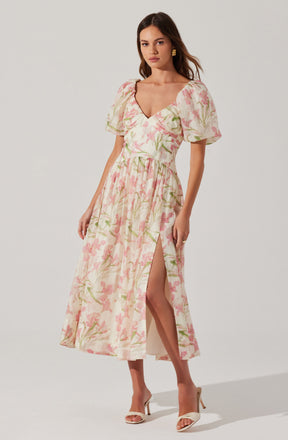 A woman is wearing a long, flowy dress with a pastel pink and green floral print. The dress has a deep V neckline, puffed short sleeves, and a high slit on one side that reveals her leg. She is also wearing beige high-heeled sandals and gold earrings.