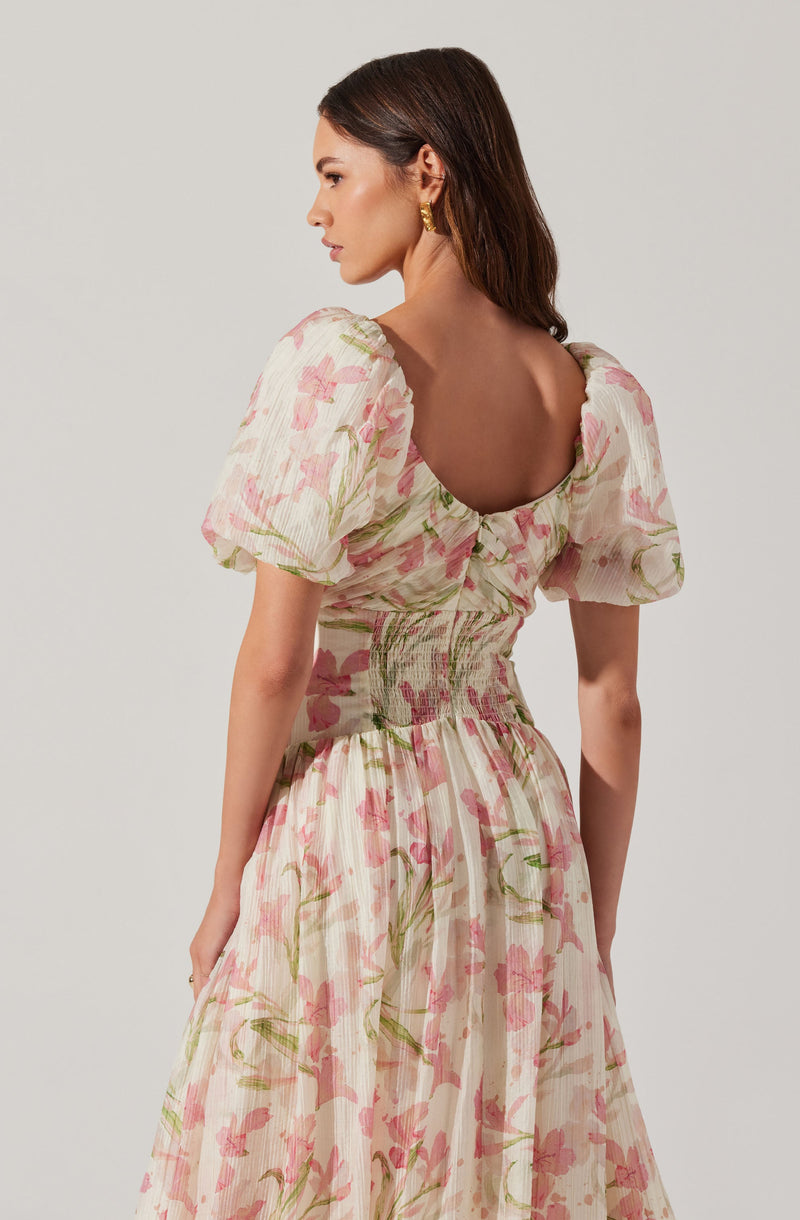 A woman is wearing a light-colored dress with a floral pattern in shades of pink and green. The dress features puffed short sleeves, a gathered waist with shirring, and a full, flowing skirt. The back of the dress has a deep, rounded neckline.