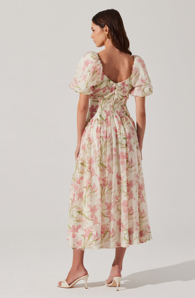 A woman is wearing a long, flowy dress with a floral pattern. The dress features puffed short sleeves, a fitted bodice with smocking detail, and a skirt that reaches mid-calf. The fabric has a light background with pink and green floral prints. She is also wearing beige high-heeled sandals.