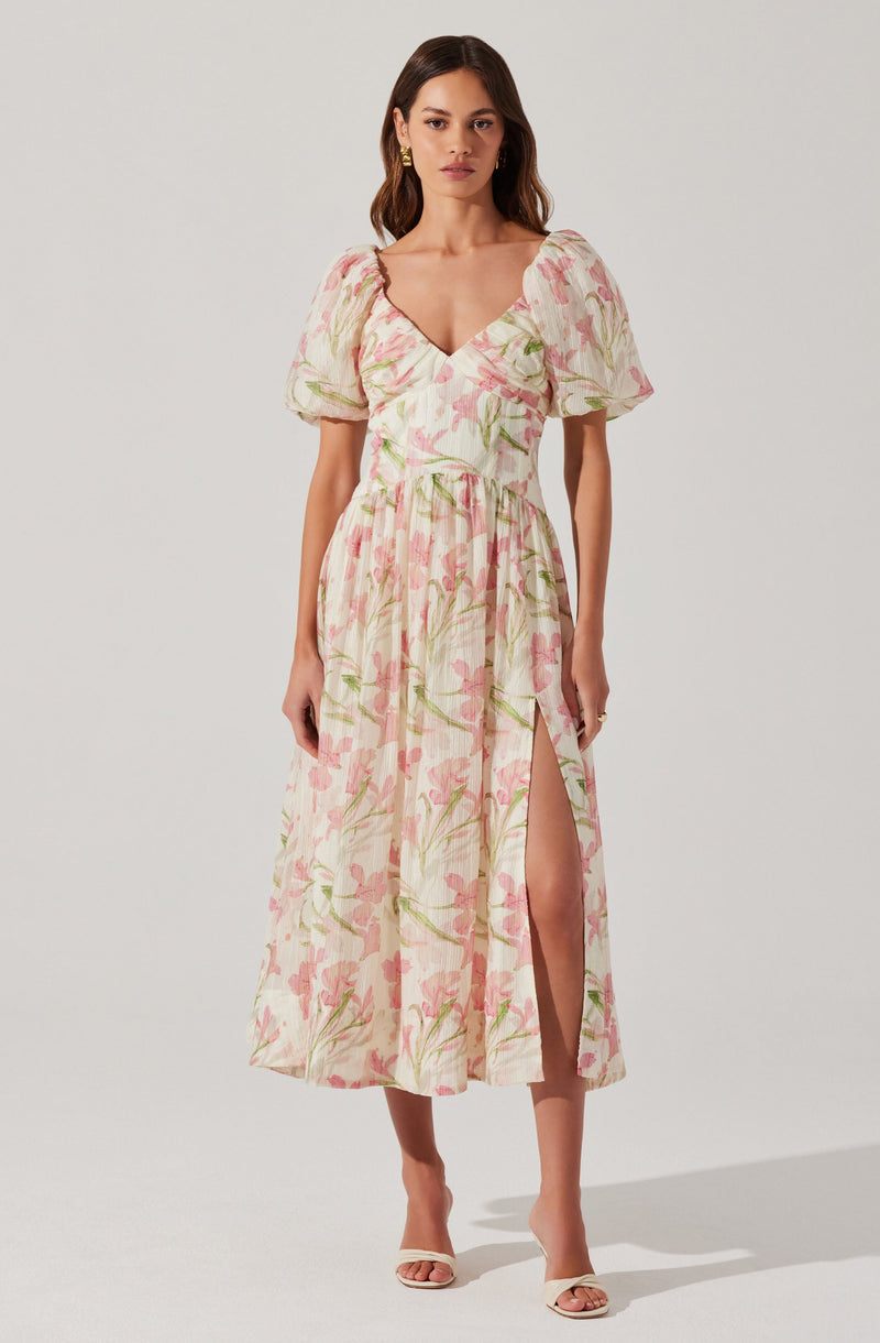 A woman is wearing a long, flowy dress with a floral pattern featuring pink and green colors on a light background. The dress has short, puffed sleeves and a deep V-neckline. It also has a high slit on one side, revealing her leg.