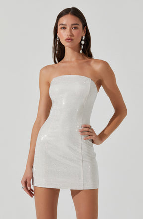 A woman is wearing a strapless, fitted mini dress made of a shiny, white fabric that appears to be embellished with small, sparkling beads or sequins. The dress is sleek and form-fitting, ending high on the thighs. She has long dark hair and is accessorized with large, dangling earrings.