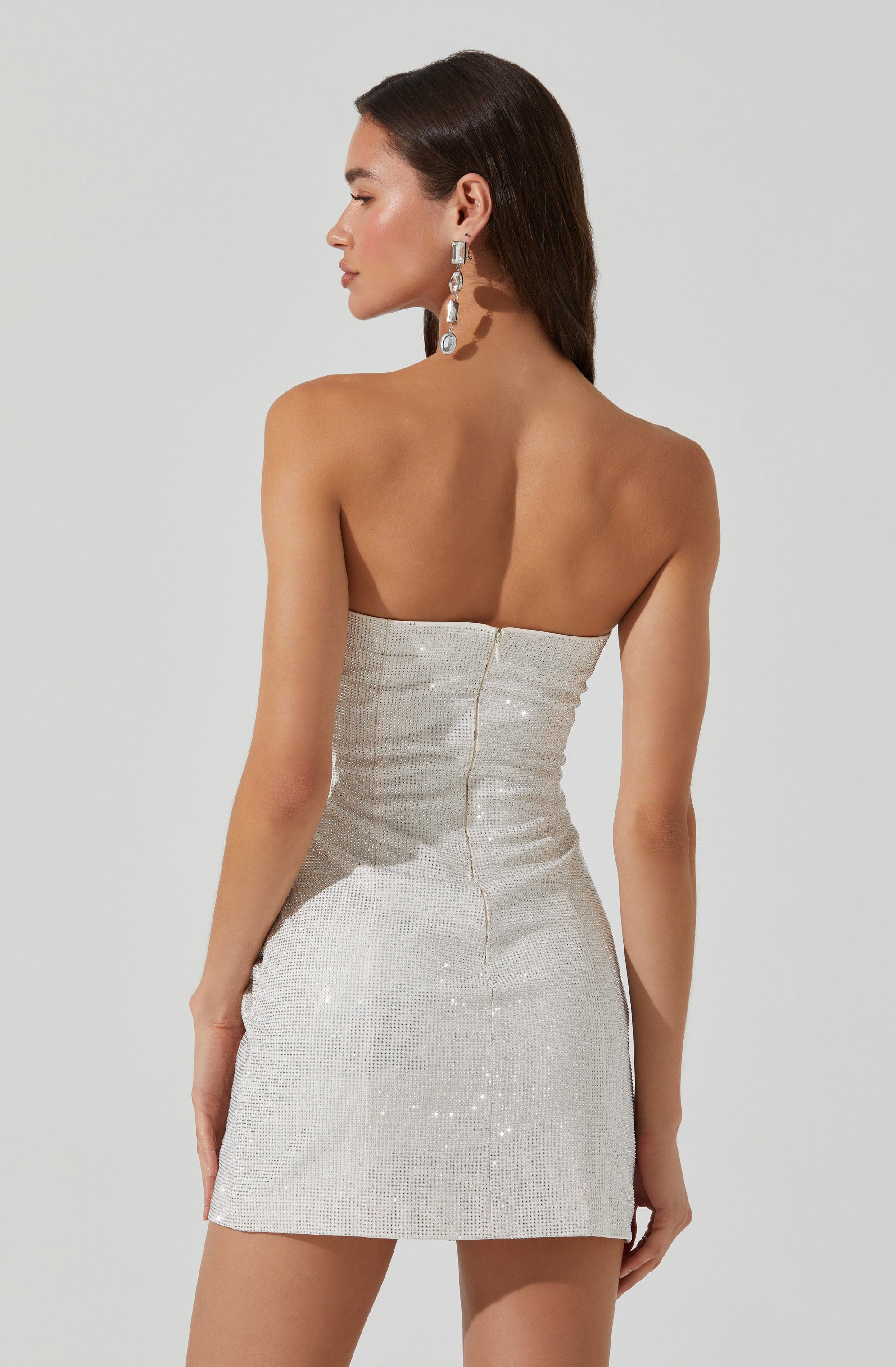 A woman is wearing a strapless, form-fitting mini dress made of a shimmering, metallic fabric with a subtle grid pattern. She has long, dark hair and is accessorized with large, dangling earrings.