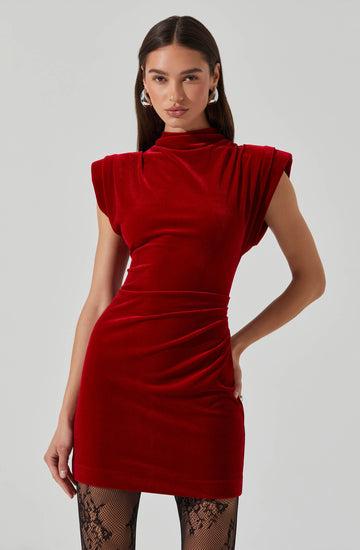 A woman is wearing a form-fitting, sleeveless red velvet dress with structured shoulder details. The dress has a high neckline and is ruched at the waist, ending above the knees. She pairs it with black patterned tights.