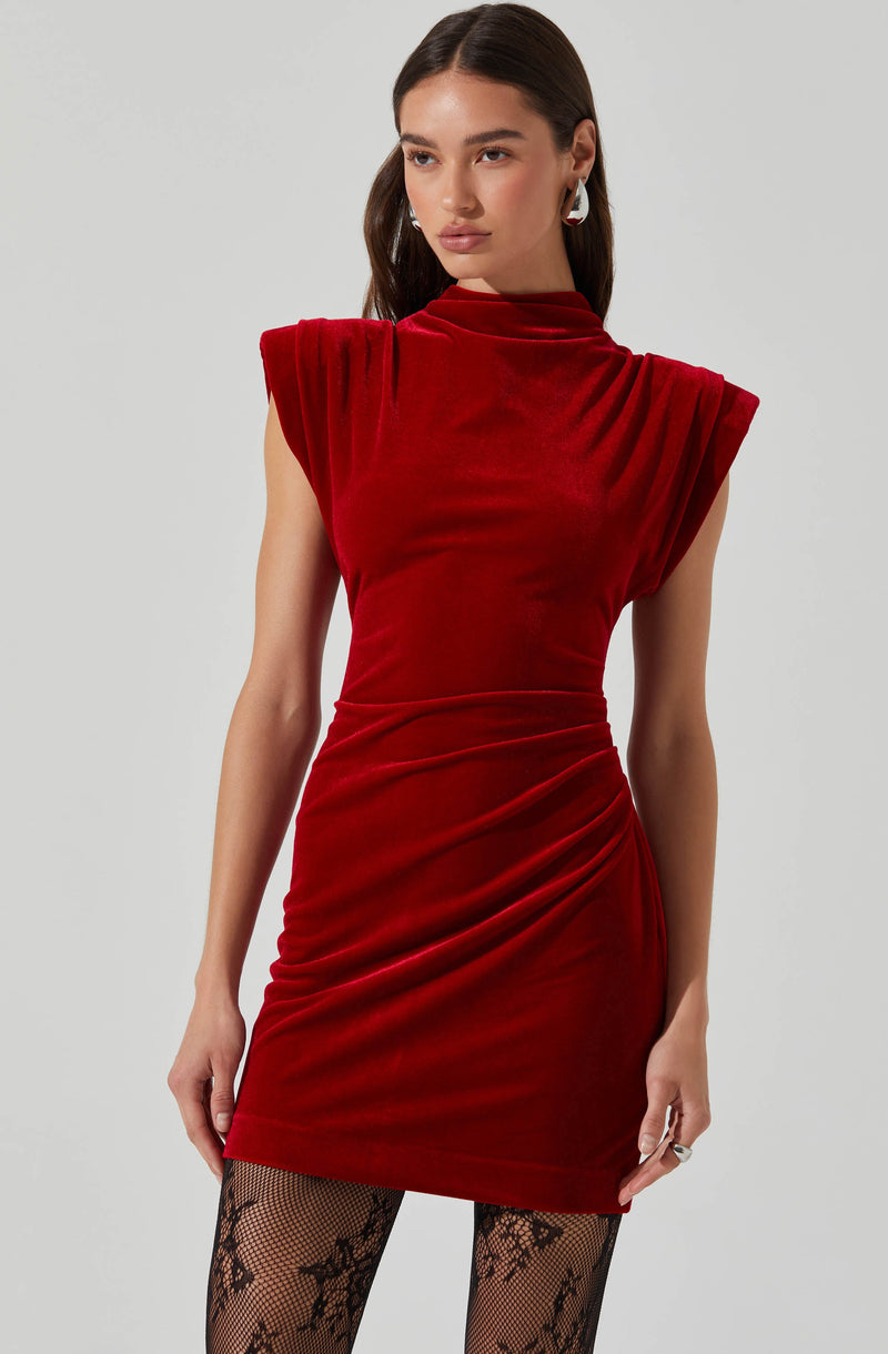 A woman is wearing a sleeveless, high-neck, fitted red velvet dress with gathered and draped details at the waist and hips. She has paired the dress with black fishnet tights featuring a floral pattern and is accessorized with a silver ring on her finger and white geometric earrings.