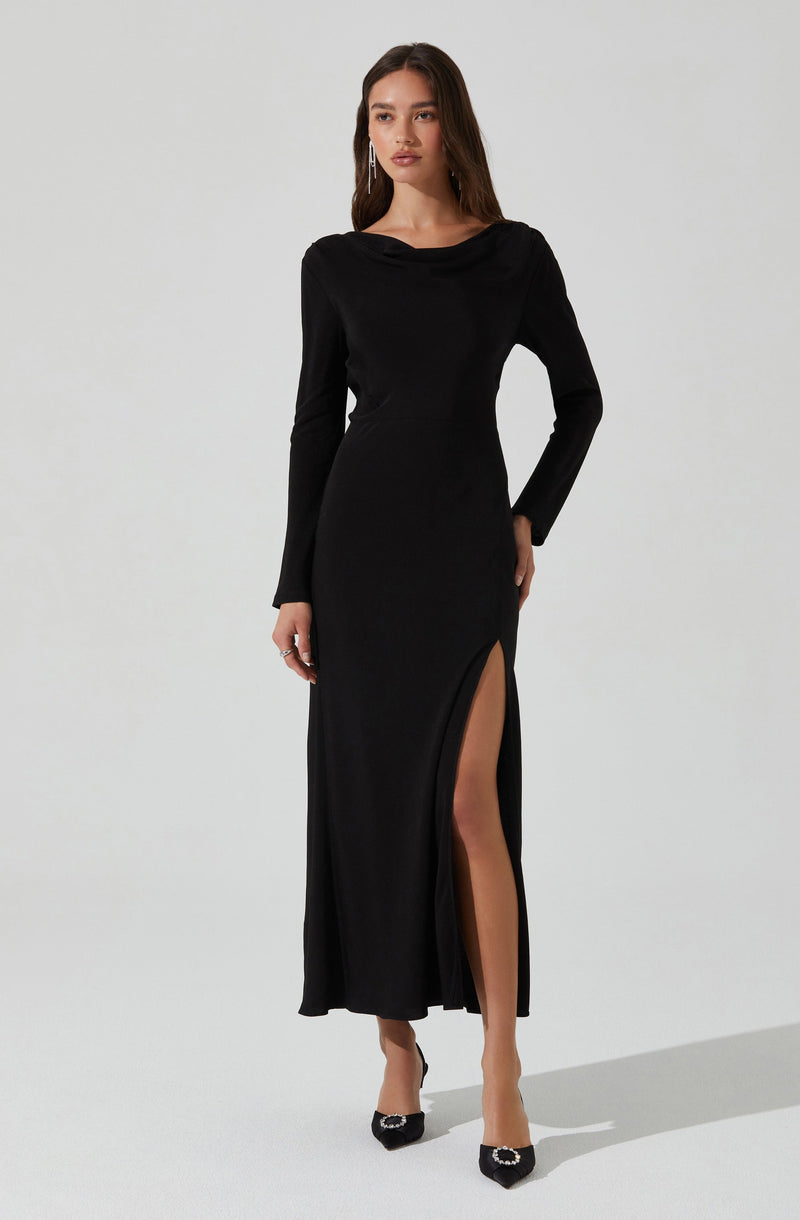 A woman is wearing a long black dress with long sleeves and a draped neckline. The dress features a thigh-high slit on the left side, revealing her leg and black high-heeled shoes with embellishments.