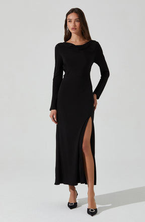 A woman is wearing a long black dress with long sleeves and a draped neckline. The dress features a thigh-high slit on the left side, revealing her leg and black high-heeled shoes with embellishments.