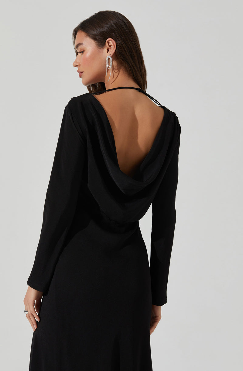 A woman is wearing a black dress with a deep open back and a hood. The dress has long sleeves and a fitted waist. She accessorizes with large, oval-shaped hoop earrings and a black choker necklace.
