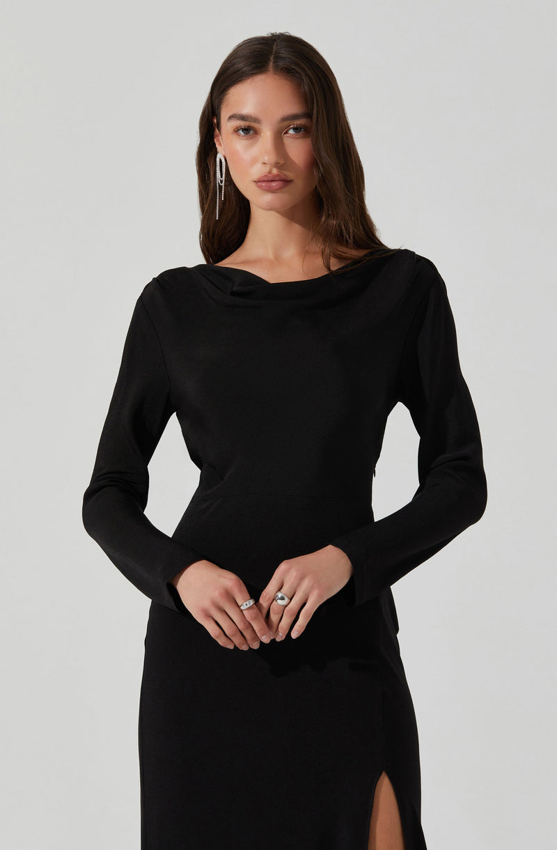 A woman is wearing a sleek, black long-sleeved dress with a subtle, gathered drape at the neckline. The dress fits closely to her body, accentuating her figure, and features a high slit on the left side. She has long, dark, wavy hair and is accessorized with large, dangling earrings and rings on her fingers.