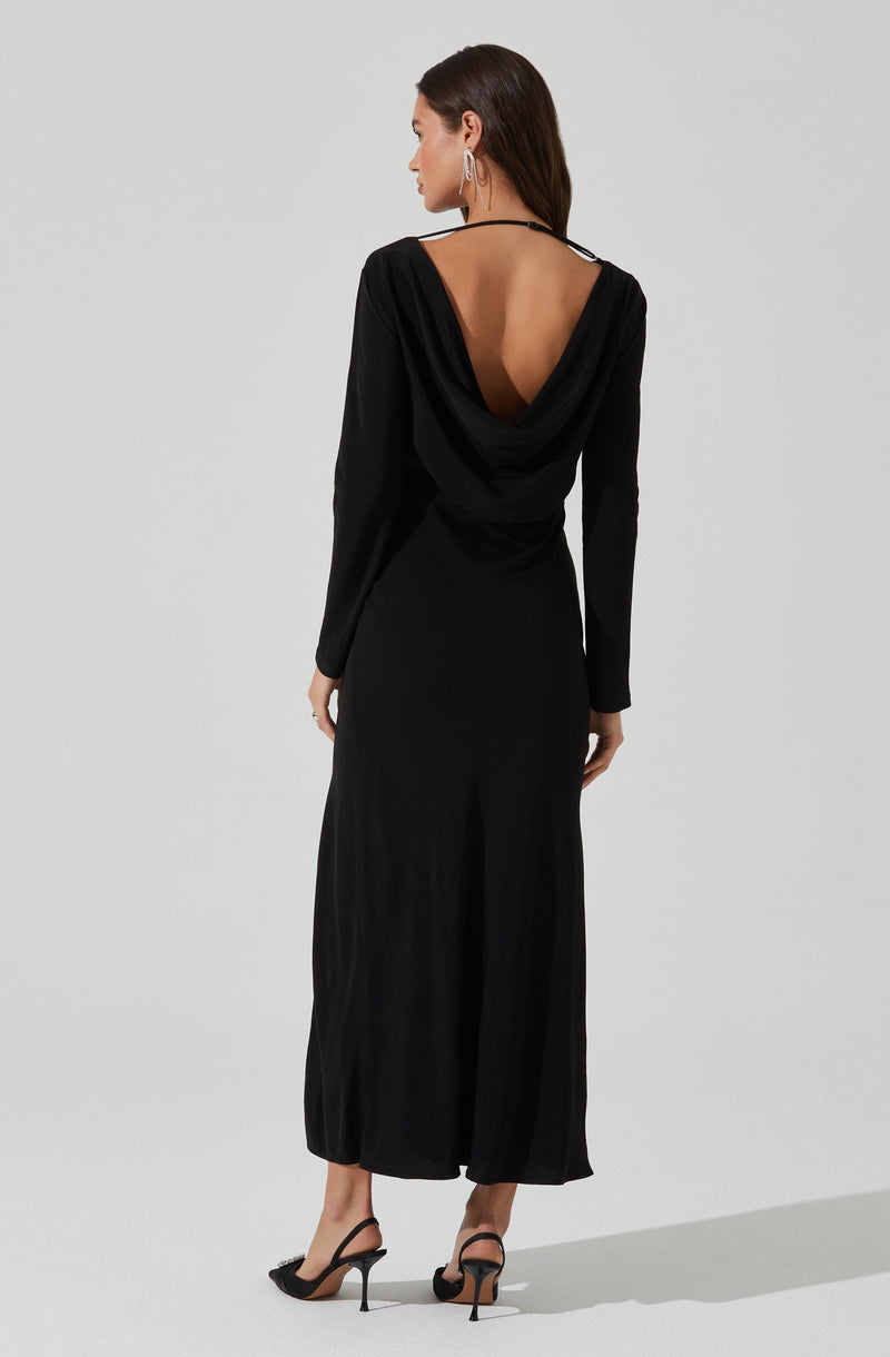 A woman is wearing a long, black dress with a deep, draped back design. The dress has long sleeves and a fitted silhouette that reaches her ankles. She is also wearing high-heeled sandals with thin straps and decorated with bows at the front.