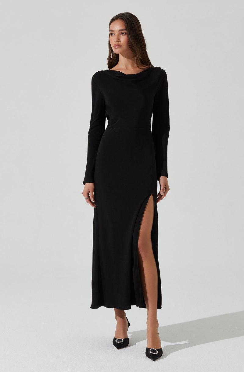 A woman is wearing a long black dress with a high slit on the left side that reveals one leg. The dress has long sleeves and a draped neckline. She is also wearing black high heels with embellishments on the toes.