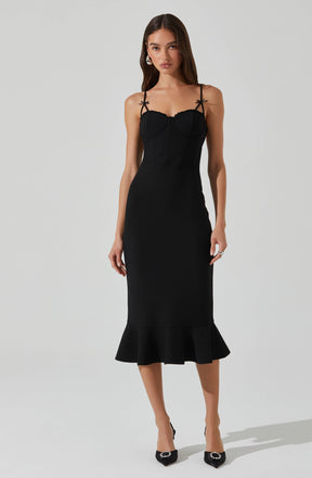 A woman wearing a black midi dress with thin, tied shoulder straps and a fitted silhouette. The dress features a ruffled hem at the bottom and appears to be made of a smooth, structured fabric. She is also wearing black high heels with embellishments and earrings.
