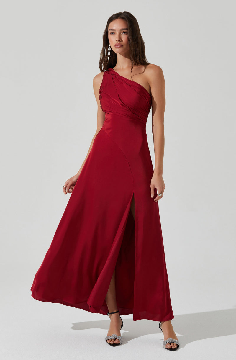 A woman is wearing a floor-length, one-shoulder red gown with ruched detailing on the bodice. The dress features a high slit in the front, revealing her legs. She pairs the gown with open-toe high heels and statement earrings.