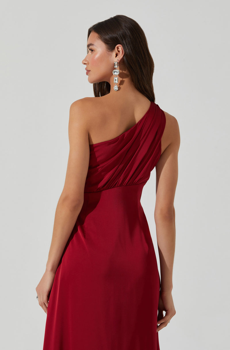 A woman is wearing a deep red, one-shoulder evening gown with pleated fabric on the bodice. The dress has a fitted waist and flows down smoothly. She is also wearing large, dangling, silver and gemstone earrings.