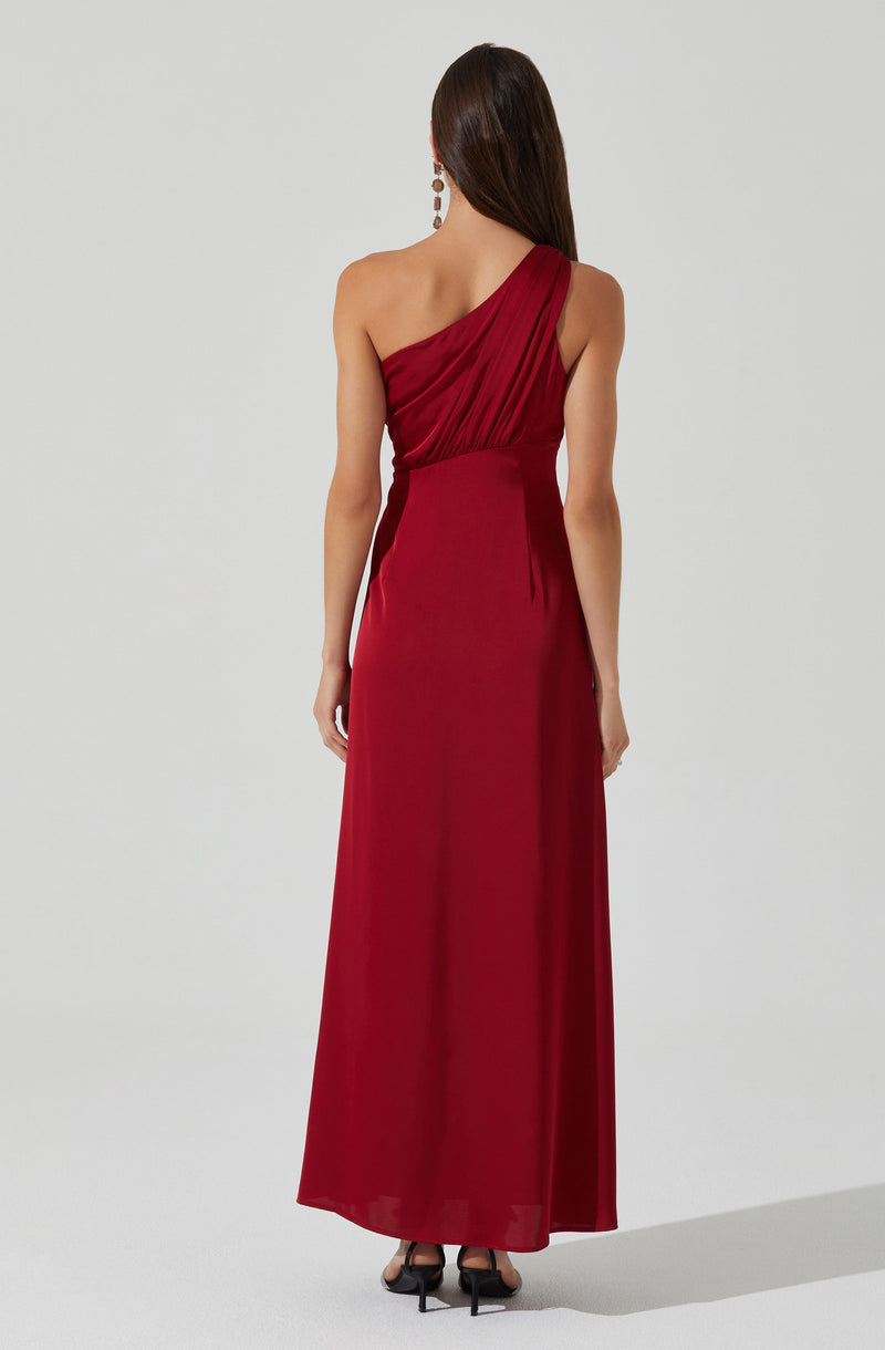 A woman is wearing a floor-length, deep red evening gown with an asymmetrical one-shoulder design. The dress features pleated fabric across the bodice and flows smoothly down to the hem. She accessorized with long, dangling earrings.