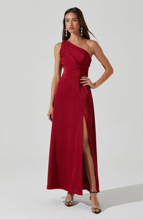 A woman is wearing a long, elegant, strapless red dress. The dress features a gathered, ruched bodice on one side and a high slit on the front left side, revealing her leg. She accessorizes with large, dangling earrings and open-toed heels with ankle straps.