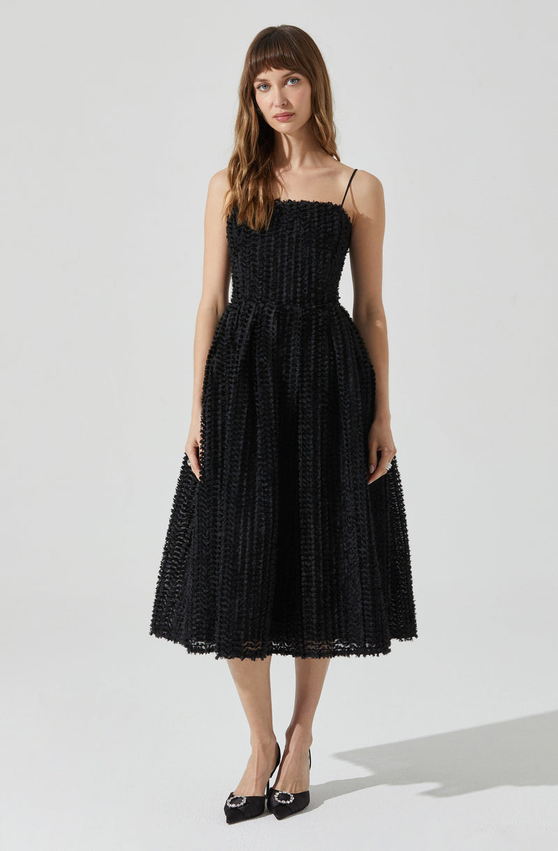 A woman is wearing a black dress with a textured, button-like pattern. The dress has thin spaghetti straps, a fitted bodice, and a full, midi-length skirt. She is also wearing black high heels with embellishments on the toes.