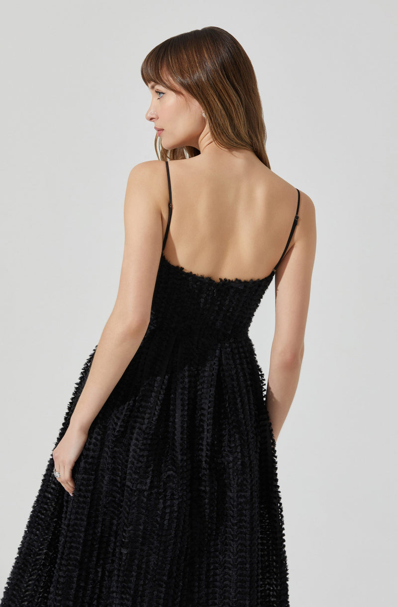 A woman is wearing a black, textured, floor-length dress with a fitted bodice. The dress features thin spaghetti straps over her shoulders. The fabric appears to have a subtle sheen and a vertically ridged or striped pattern, giving it a rich, tactile look.