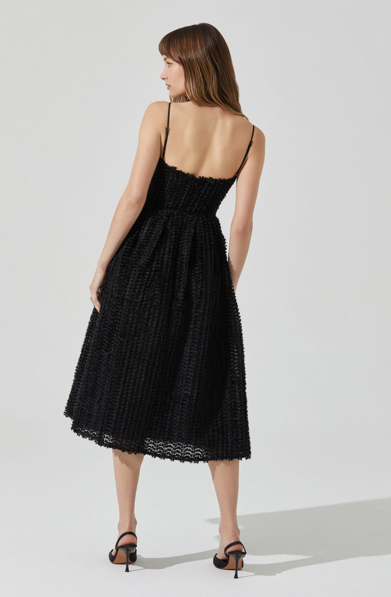 A woman is wearing a black, textured, sleeveless midi dress with thin spaghetti straps. The dress has a fitted bodice and a full skirt, featuring a woven or crochet-like pattern. She is also wearing black high-heeled sandals.