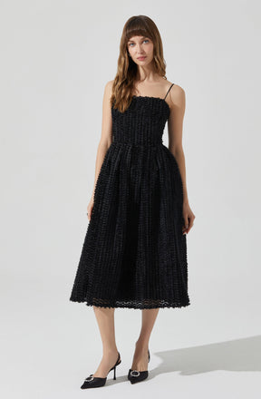 A woman is wearing a black, textured, sleeveless dress with thin spaghetti straps. The dress has a fitted bodice and a full, midi-length skirt with a waffle-like pattern. She pairs the dress with black pointed-toe high heels that have decorative embellishments on the front.