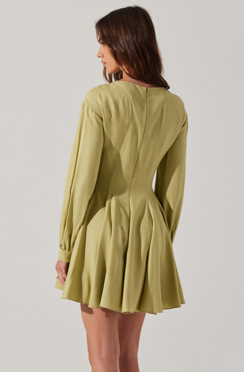 A woman is wearing a light olive-green dress with long sleeves. The dress features a fitted waist with pleats that create a flared, skirt-like bottom. The fabric appears to be smooth and slightly shiny. The back of the dress has a center seam running from the neckline to the waist.