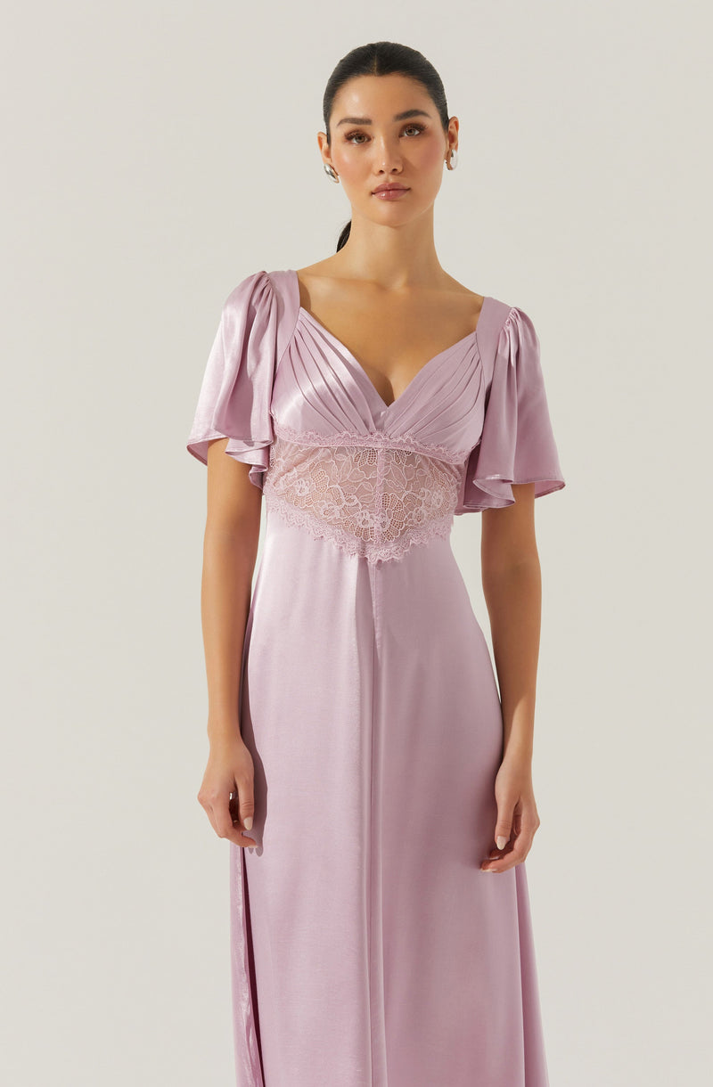 A woman is wearing a floor-length, pastel pink dress with short, puffed sleeves made of a shiny, satin-like fabric. The dress features a V-neckline and a lace band around the waist with floral embroidery, creating a fitted silhouette.