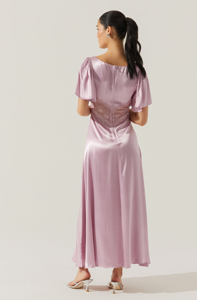 A woman is wearing a long, silky, light pink satin gown with short puffed sleeves. The dress features a lace detail around the waist and a fitted bodice, flowing down to the floor. She is also wearing open-toe heels with a small heel. Her dark hair is styled in a high ponytail.