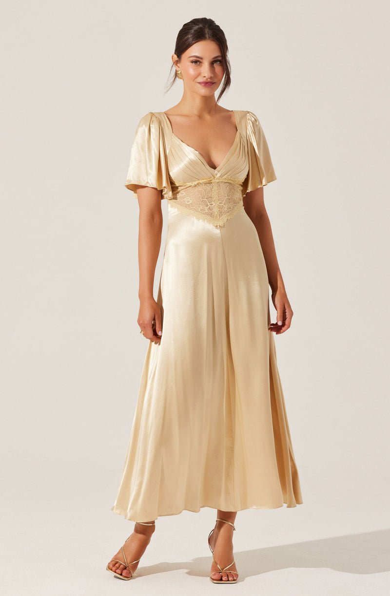 A woman is wearing an elegant, floor-length, champagne-colored satin gown with a deep V neckline. The dress features short, puffed sleeves and an embroidered or lace bodice area that extends to the waist, creating a fitted look. She is also wearing nude high-heeled sandals.