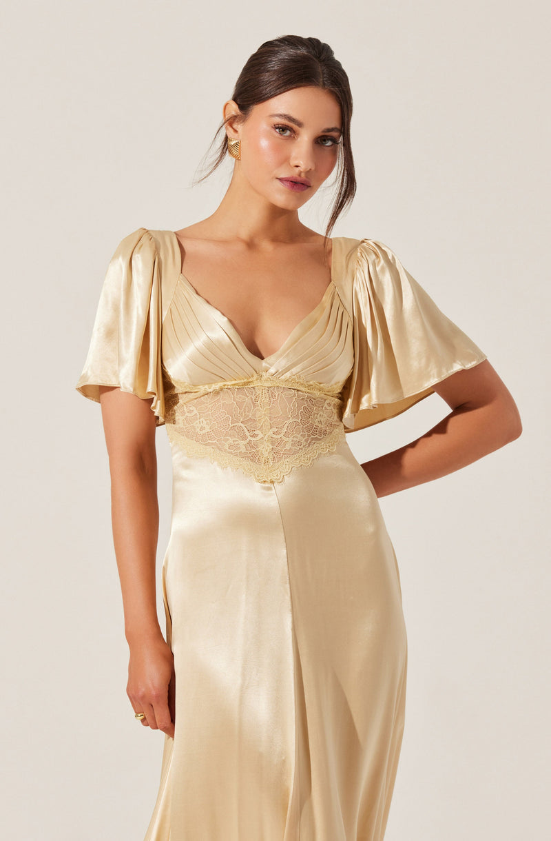 A woman is wearing a satin gown in a champagne color. The dress features short, puffed sleeves and a fitted bodice with pleated fabric and lace detailing at the waist. The skirt is smooth and flowing, with a subtle slit at the front.