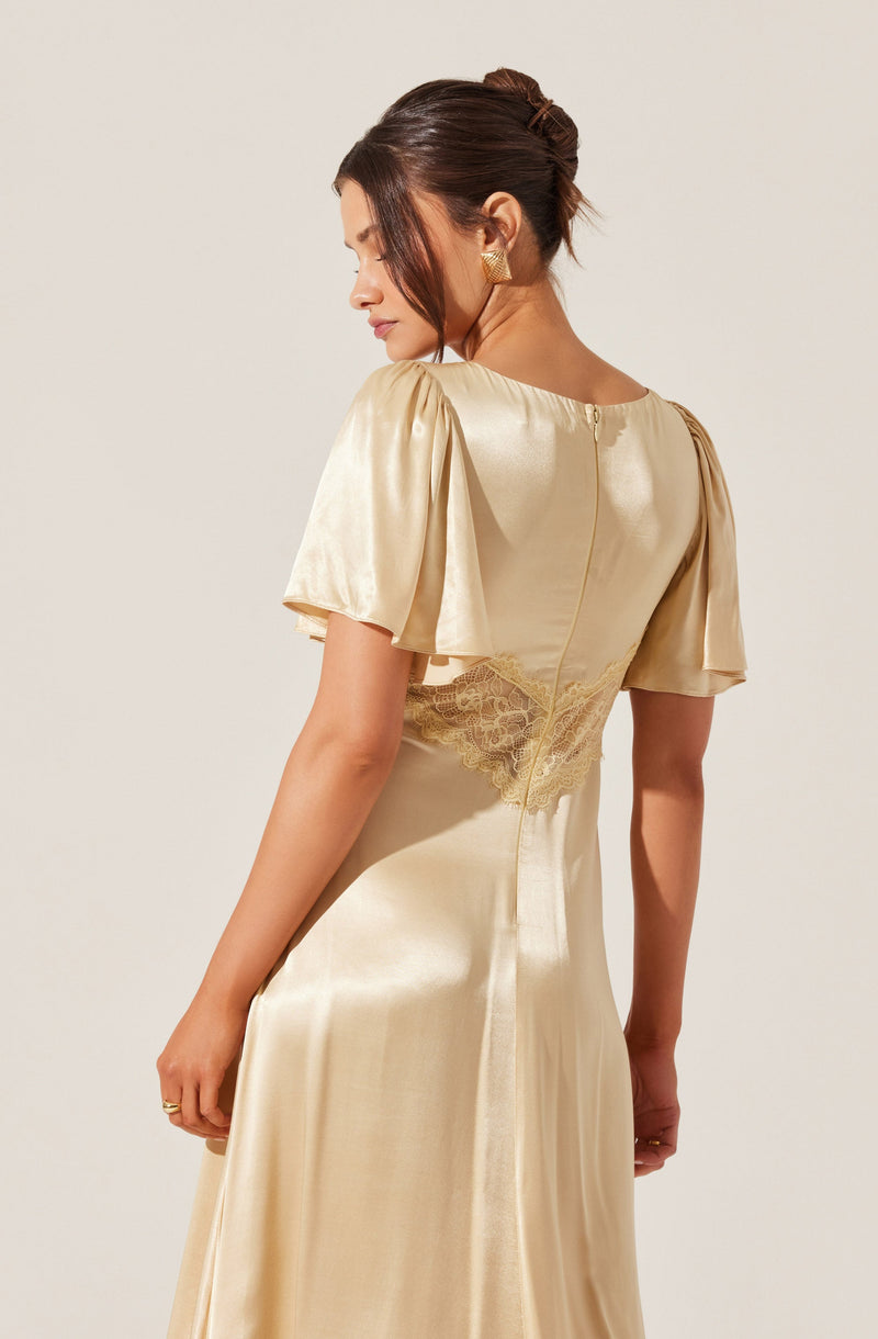 A woman is wearing a silky, champagne-colored satin dress with puffed short sleeves. The dress features a form-fitting design with lace detailing around the waist and a back zipper closure. She is also wearing large, triangular gold earrings.