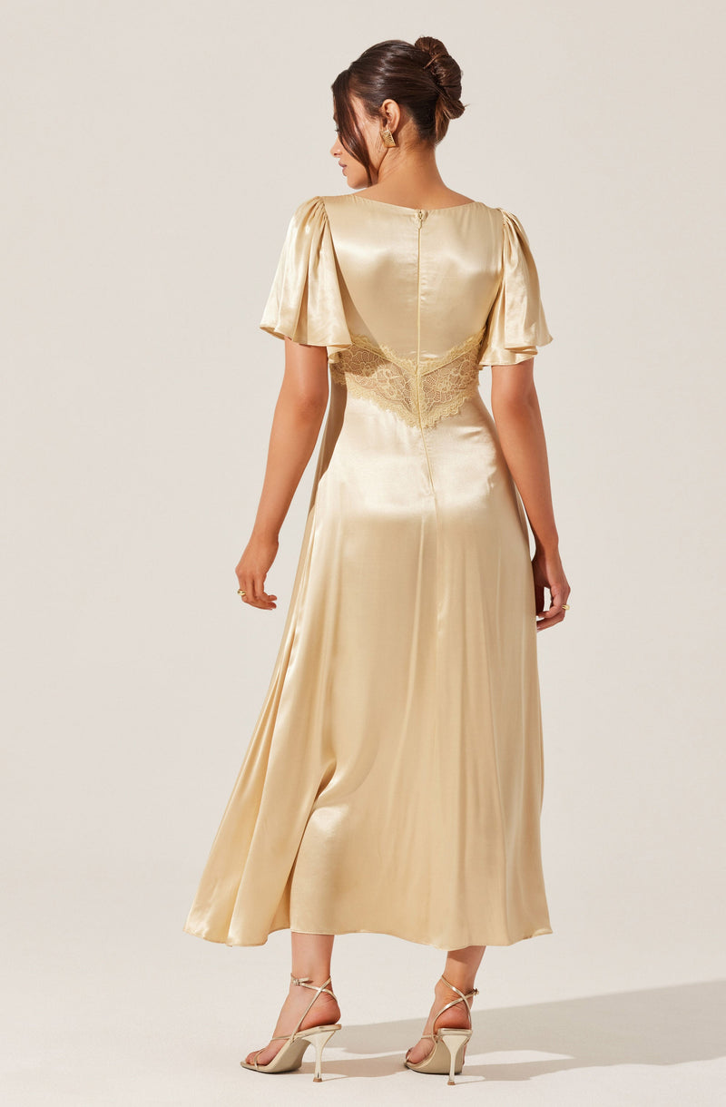 A woman is wearing a long, elegant, satin gown in a champagne color. The dress features puffed short sleeves and a fitted waist with lace detailing at the back. The skirt flows down to her ankles with a slight sheen and a slit at the back. She is also wearing strappy high-heeled sandals.