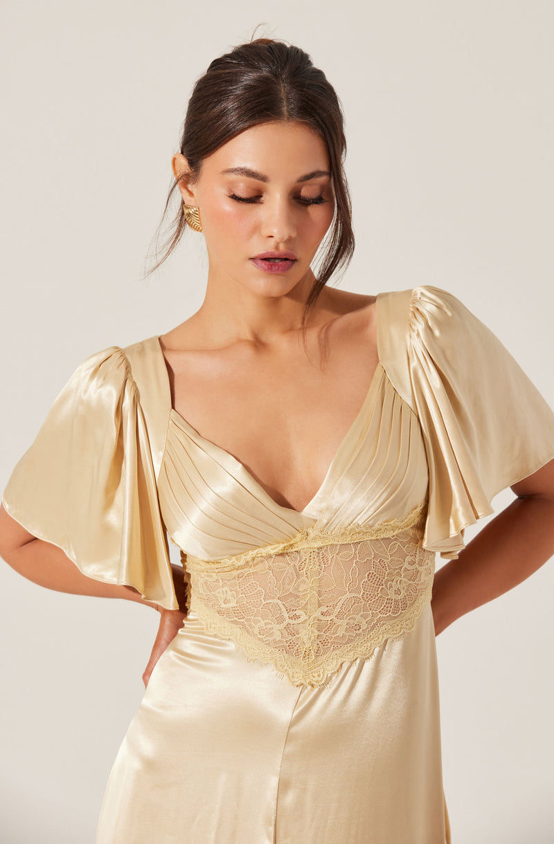 A woman is wearing a light gold satin dress with puffed short sleeves that have draped, gathered fabric details. The dress has a V-neckline and features a lace panel at the waist, adding a delicate touch to the elegant fabric. She is also wearing gold earrings.