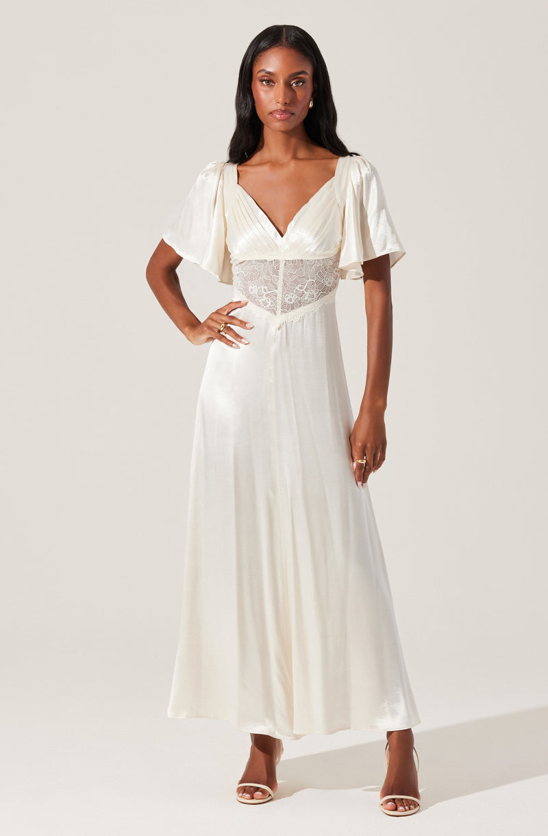 A woman is wearing a long, elegant white dress with short, puffed sleeves made of a flowing, silky fabric. The dress features a V-neckline and a sheer lace panel around the waist, adding a delicate touch. She is also wearing high-heeled shoes and accessorized with rings and earrings.