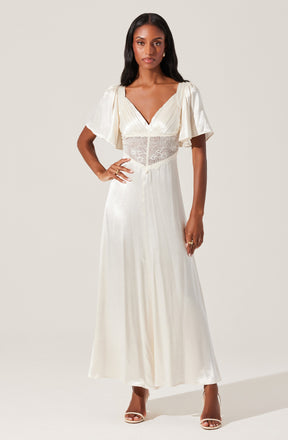 A woman is wearing a long, elegant white dress with short, puffed sleeves made of a flowing, silky fabric. The dress features a V-neckline and a sheer lace panel around the waist, adding a delicate touch. She is also wearing high-heeled shoes and accessorized with rings and earrings.