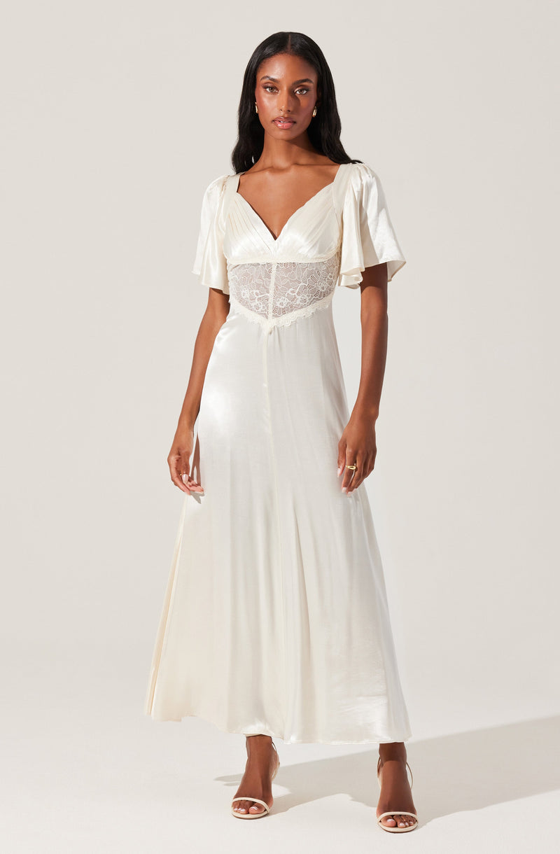 A woman is wearing a long, flowing white dress with a V-neckline and short, loose sleeves. The top part of the dress features a sheer lace panel around the waist, adding a delicate touch. She is also wearing open-toed high-heeled sandals.