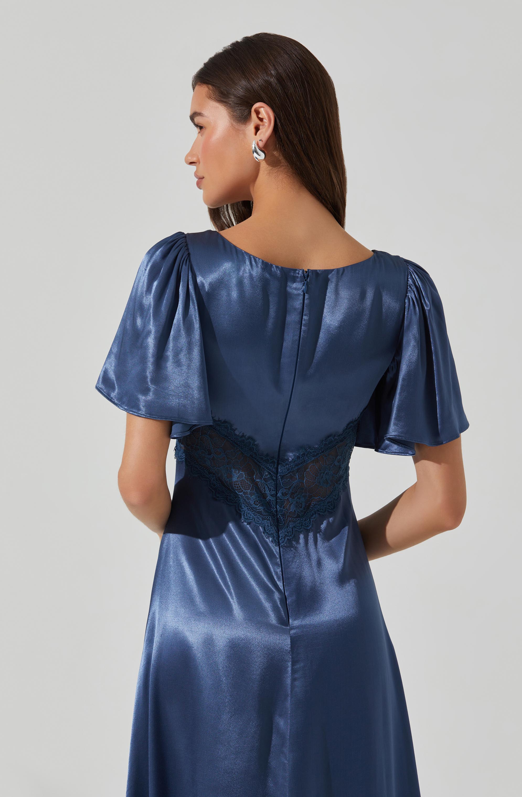 A woman is wearing a navy blue satin dress with short, puffed sleeves and a fitted waist. The dress features a lace panel at the back, adding a decorative touch. She has long, straight brown hair and is wearing silver hoop earrings.