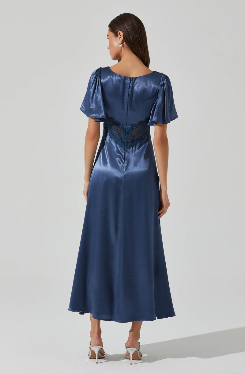 A woman is wearing a long, satin, navy blue dress with puffed short sleeves and a round neckline. The dress features a fitted waist with lace detailing and a full-length flowing skirt. She is also wearing high-heeled shoes.
