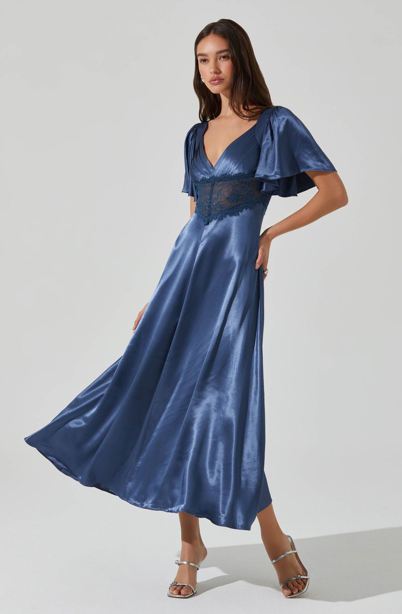 A woman is wearing a long, shiny, satin blue dress featuring a deep V neckline, puffed short sleeves with ruffles, and a waist detail with lace trim. She is also wearing high-heeled sandals.