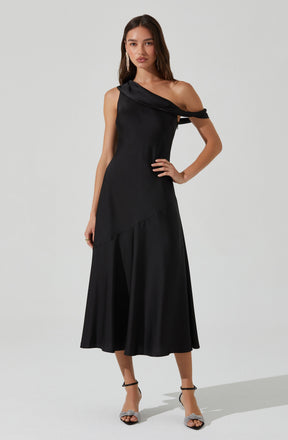 A woman is wearing a black, asymmetrical, one-shoulder dress with a draped sleeve. The dress has a fitted bodice and a flowing, slightly flared skirt that reaches mid-calf. She is also wearing black strappy high-heeled sandals with embellishments on the front straps.