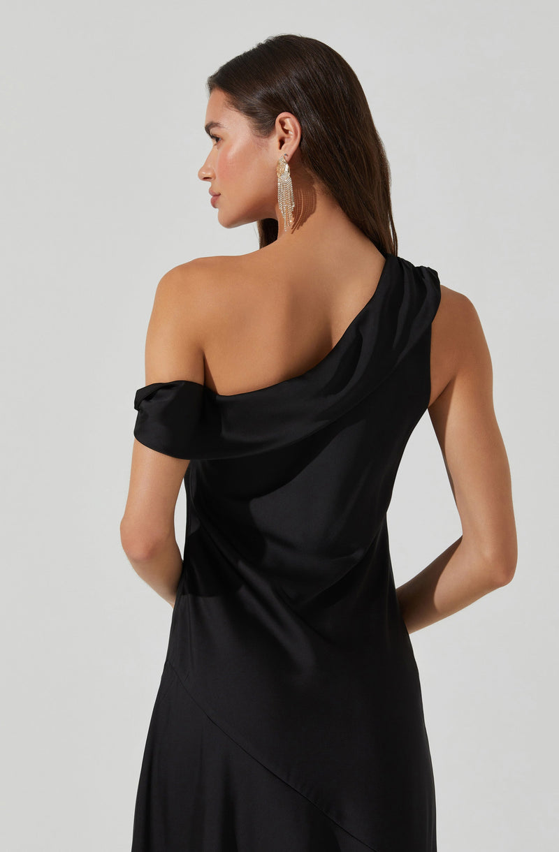 A woman is wearing a black satin dress with a single shoulder strap that drapes over her right shoulder. The dress has an asymmetrical design, with a ruched sleeve on one side and an off-the-shoulder style on the other. She is accessorized with large, dangling earrings featuring multiple strands of sparkling beads or crystals.
