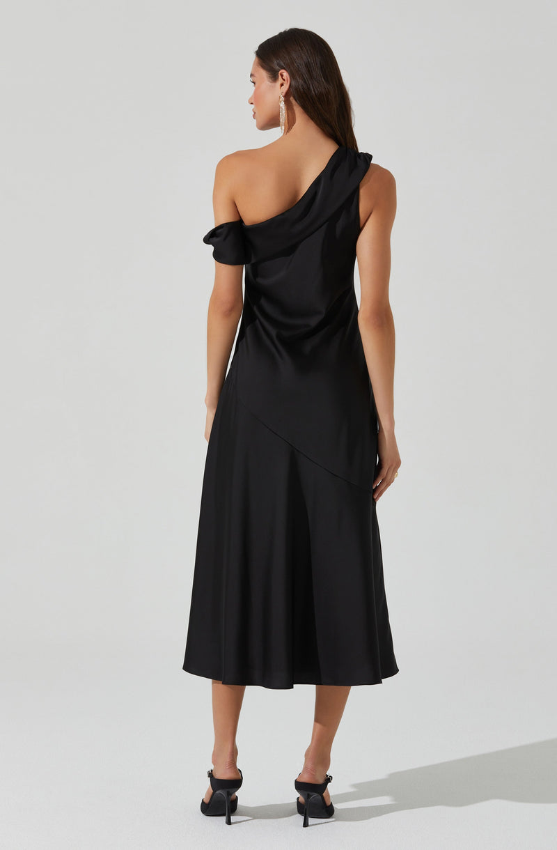 A woman is wearing a black, one-shoulder, floor-length dress with a draped neckline that creates a flowing silhouette. The dress has a unique, asymmetrical design with an off-the-shoulder sleeve on the left and a single strap on the right, giving it a sophisticated and elegant look. She is also wearing black high-heeled shoes and large, dangling earrings.