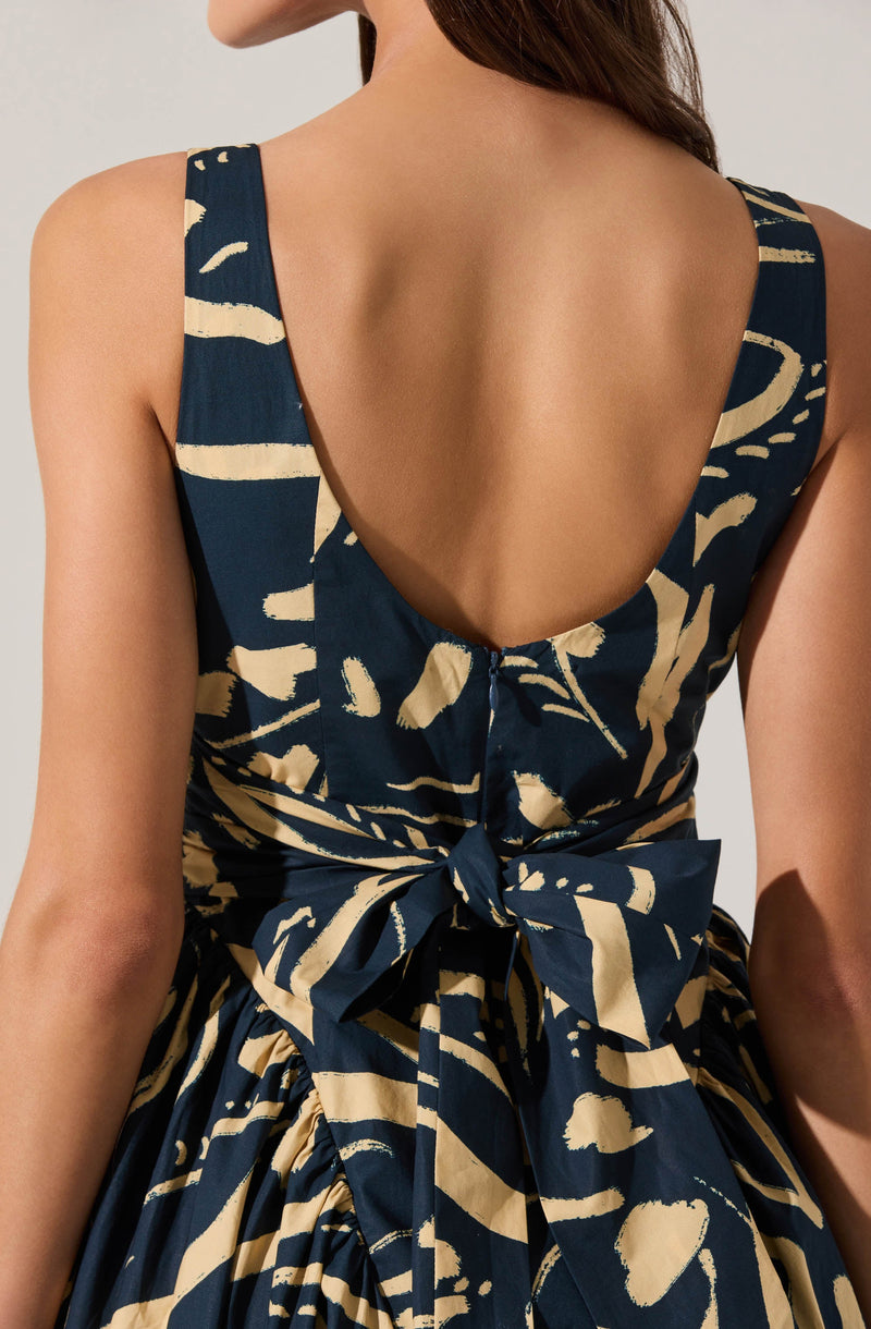 The dress features broad shoulder straps and a deep, rounded back. It has a bold, abstract pattern consisting of dark navy blue and beige brushstroke-like designs. The fabric is gathered around the waist, where it is tied into a large decorative bow at the back.